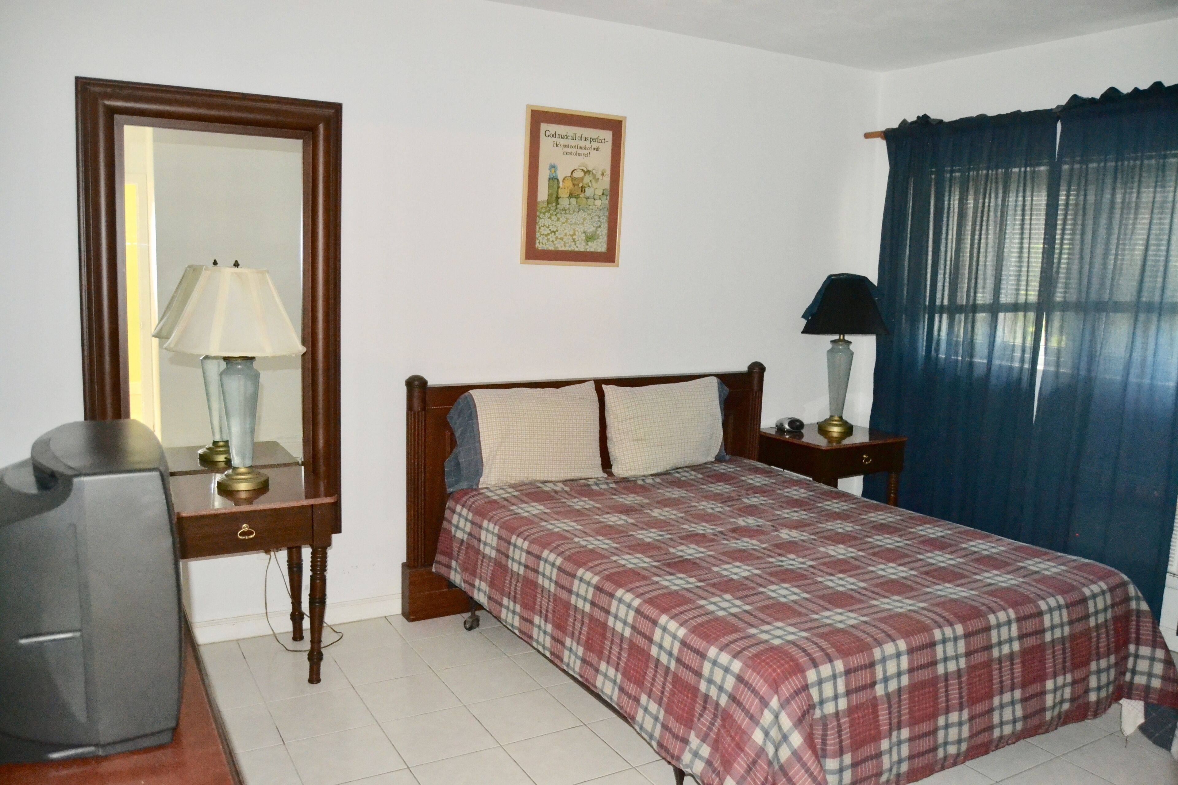 A bedroom with a bed, a desk with a lamp, a television, a framed picture on the wall, and a window with curtains.