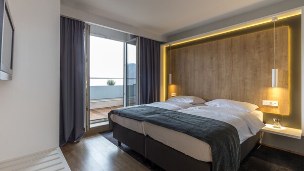 Una habitación de hotel moderna con una cama grande, una mesita de noche con lámpara, un revestimiento de pared de madera y vistas al exterior.