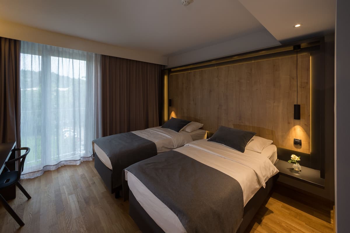 A hotel room with two beds, a desk, a chair, and a large window with curtains.
