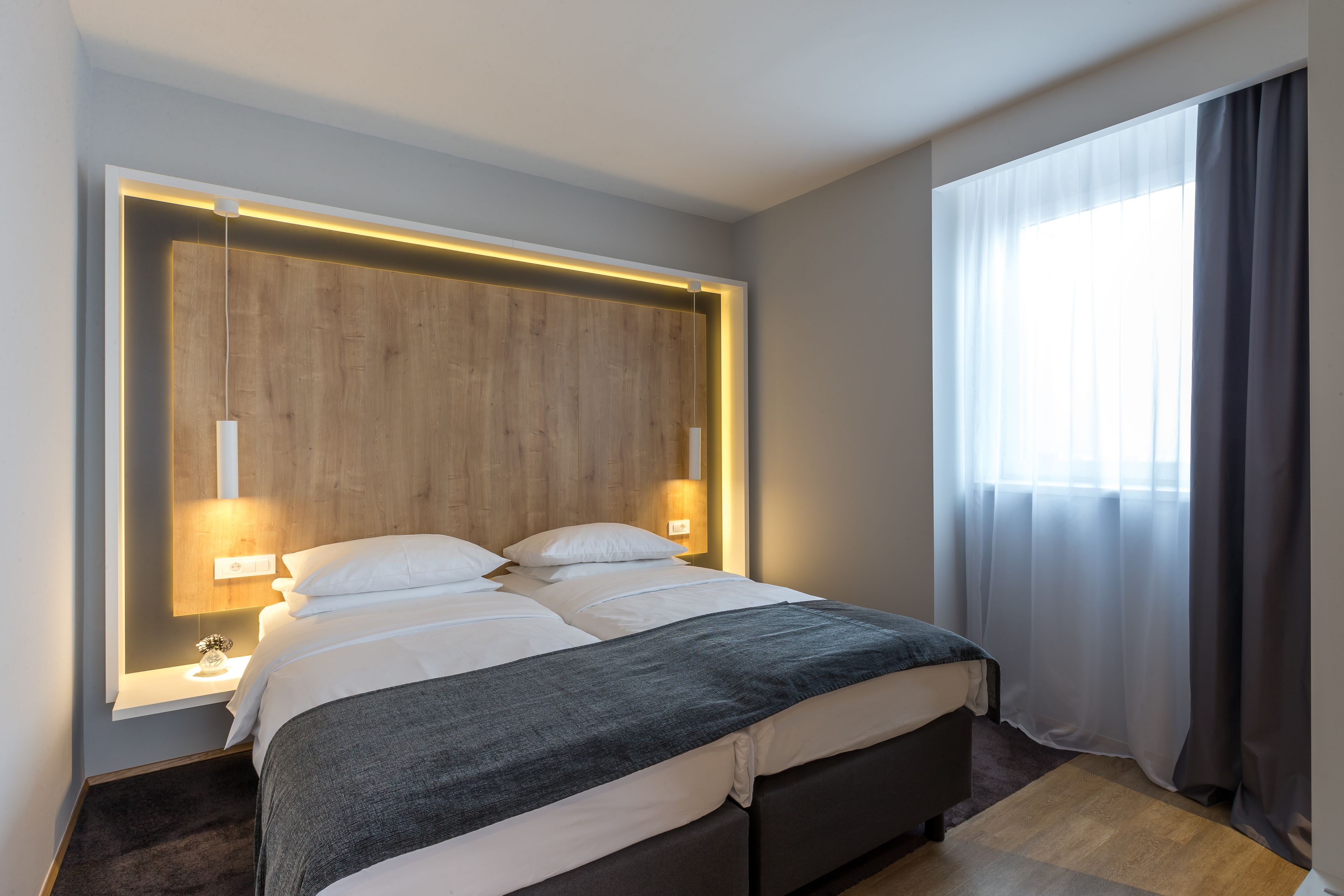 Un moderno cuarto de hotel con una cama grande, cabecera de madera, lámparas de noche y vistas al exterior a través de cortinas traslúcidas.