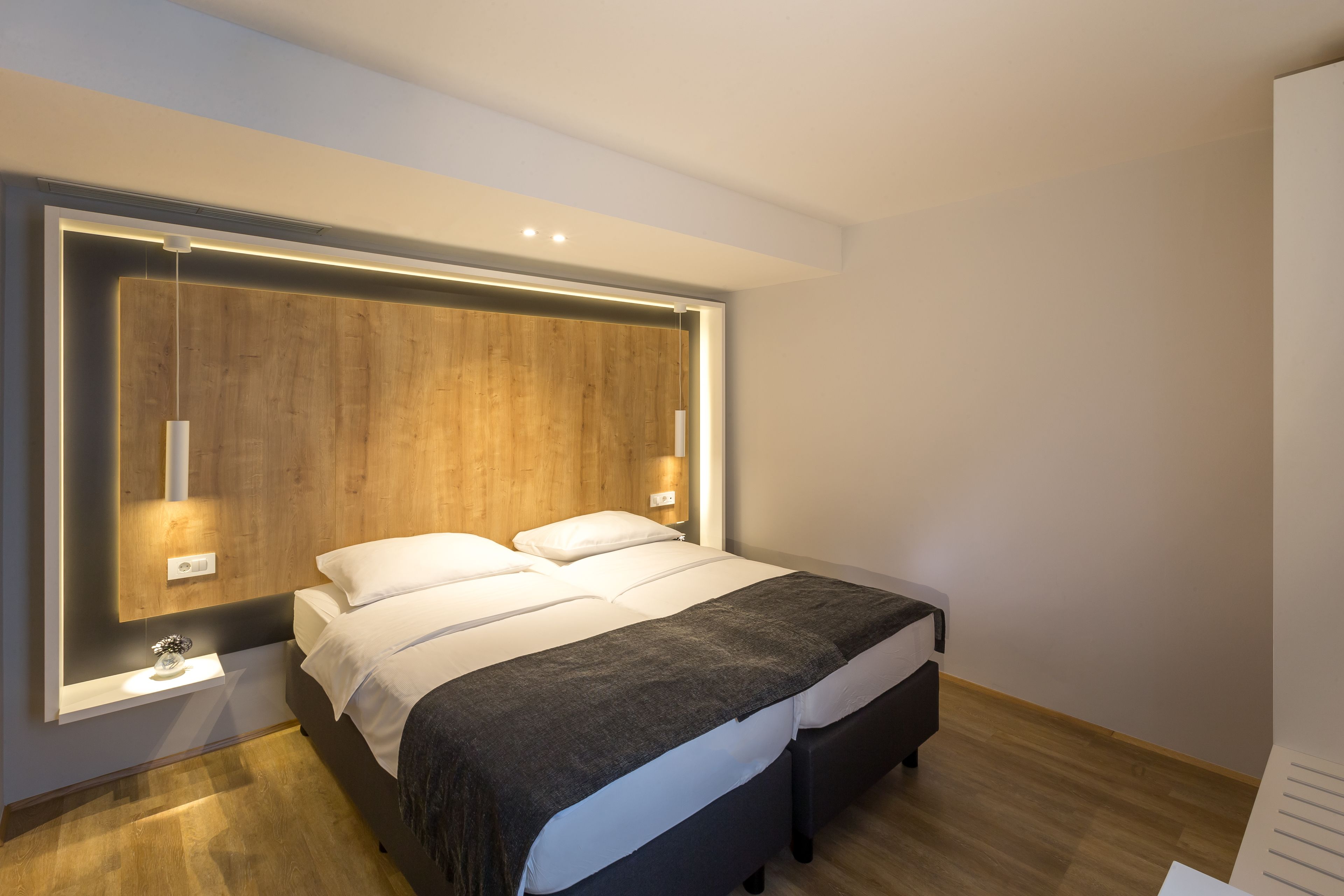 Une chambre moderne avec une tête de lit en bois, un lit recouvert de linge de maison blanc, une table de chevet et des lampes fixées au mur.