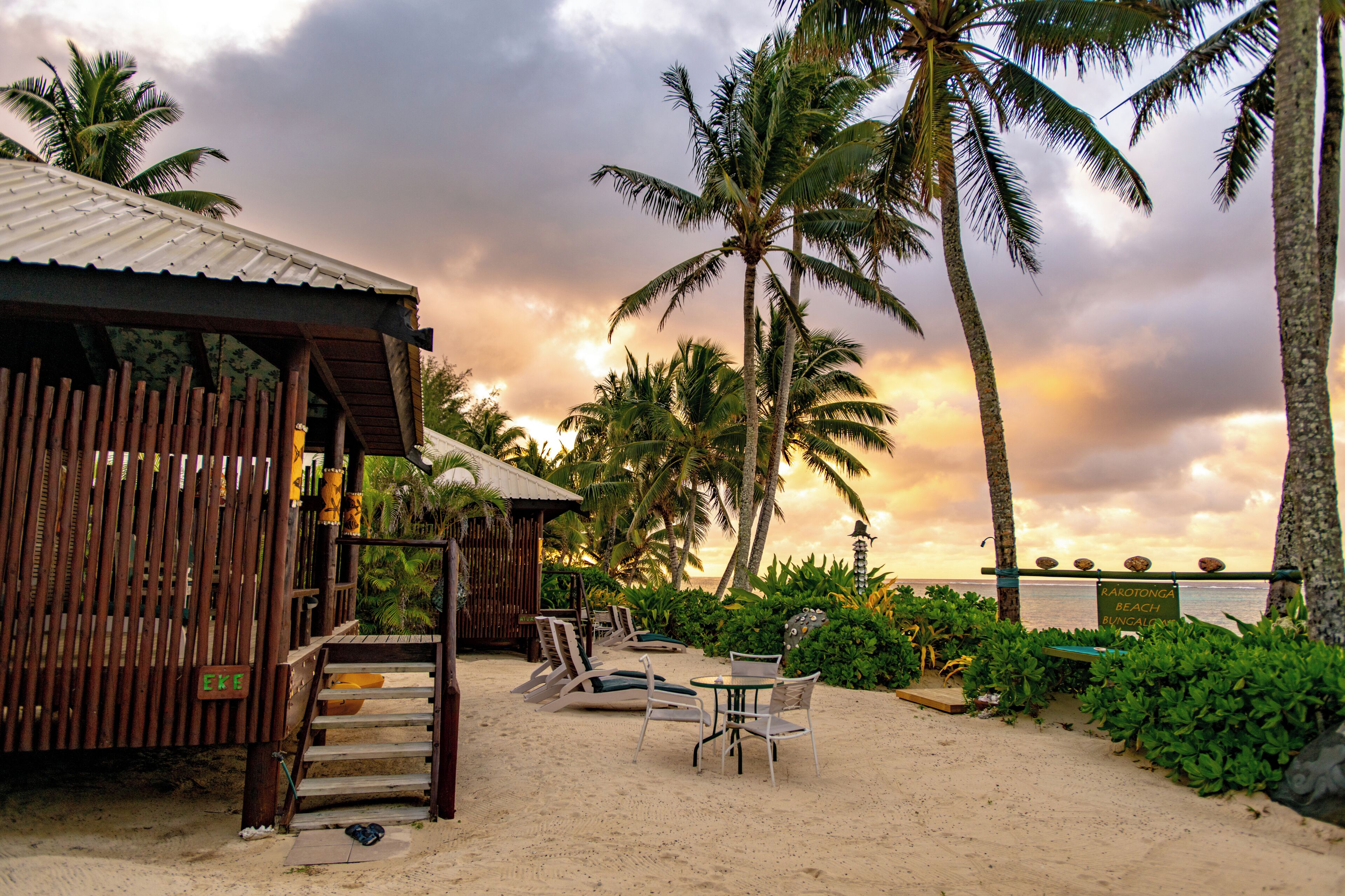 A beachside bungalow with a wooden deck, lounge chairs, and a table.