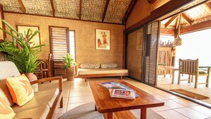 A room with a wooden bench, a sofa, a table with magazines, and a large window with blinds. - Rarotonga Beach Bungalows (Rarotonga)