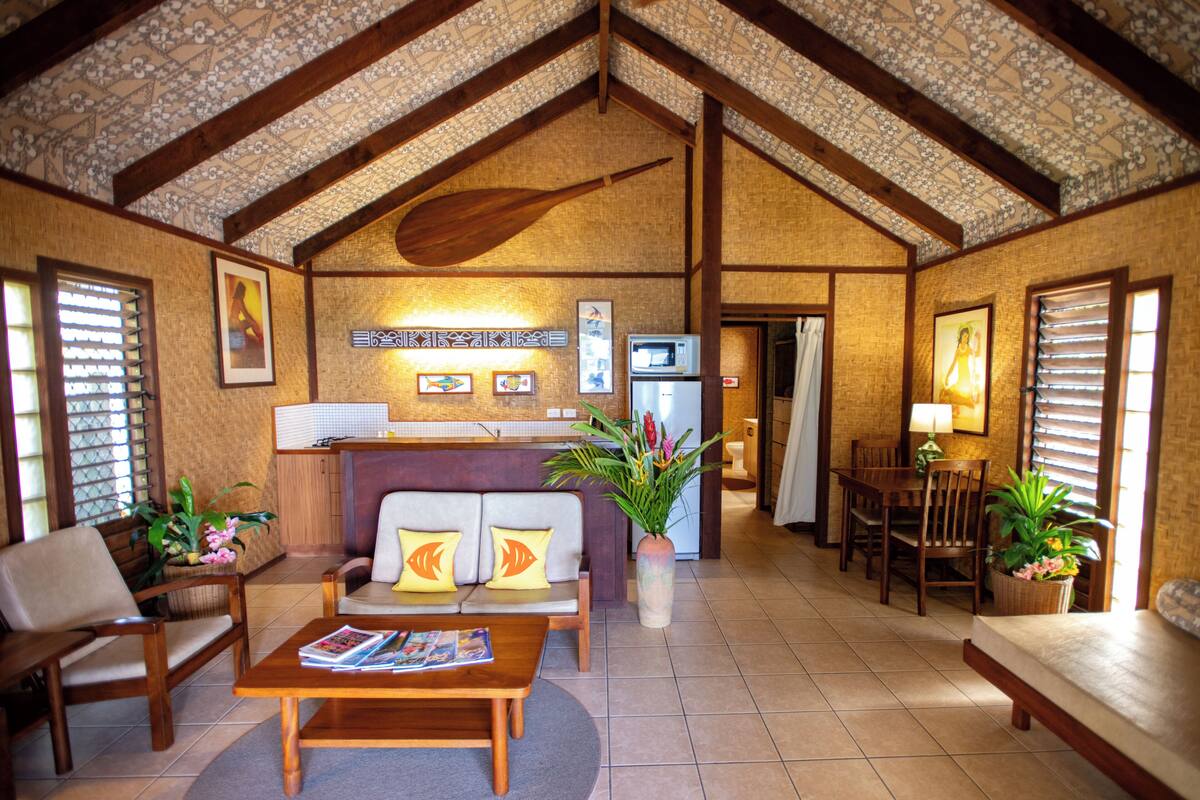 A spacious living area with a high ceiling, wooden beams, a sofa with cushions, a coffee table, a dining table with chairs, and a refrigerator.