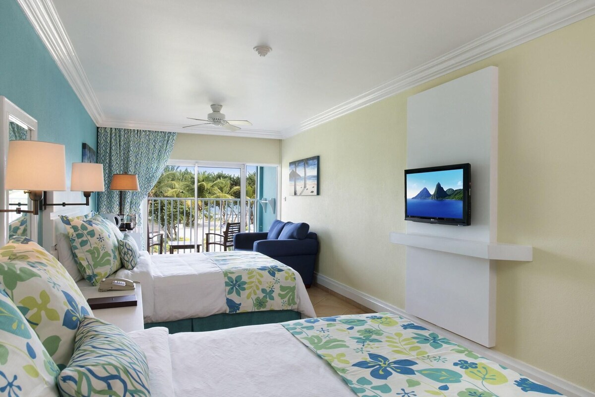 A hotel room with two beds, a TV, a ceiling fan, and a view of palm trees and mountains.