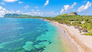 Une plage à l’eau turquoise et limpide, une rive sablonneuse, et en arrière-plan, des collines verdoyantes et une montagne.