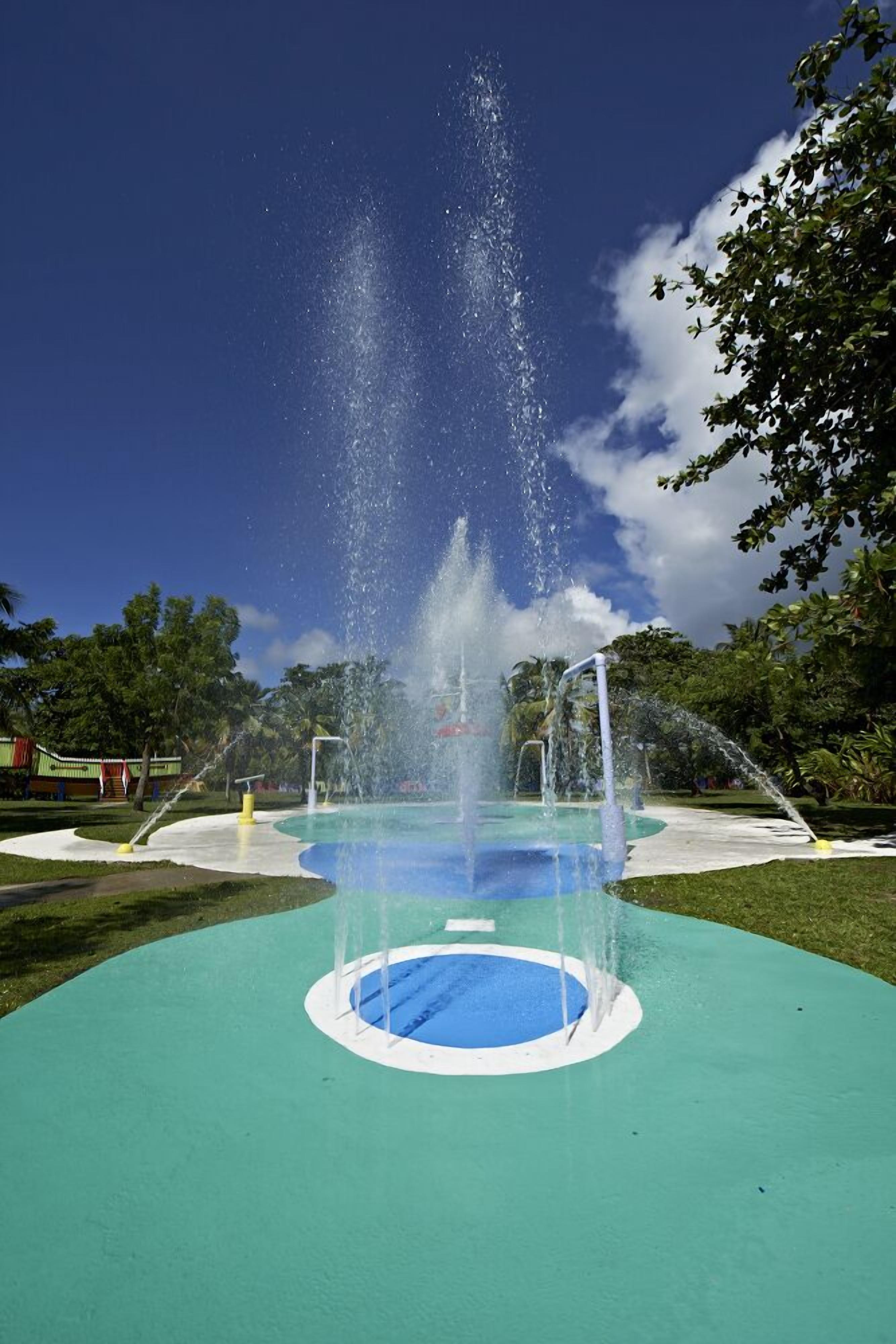 A fountain with water jets in a park with a blue circular area in the foreground and trees in the background.