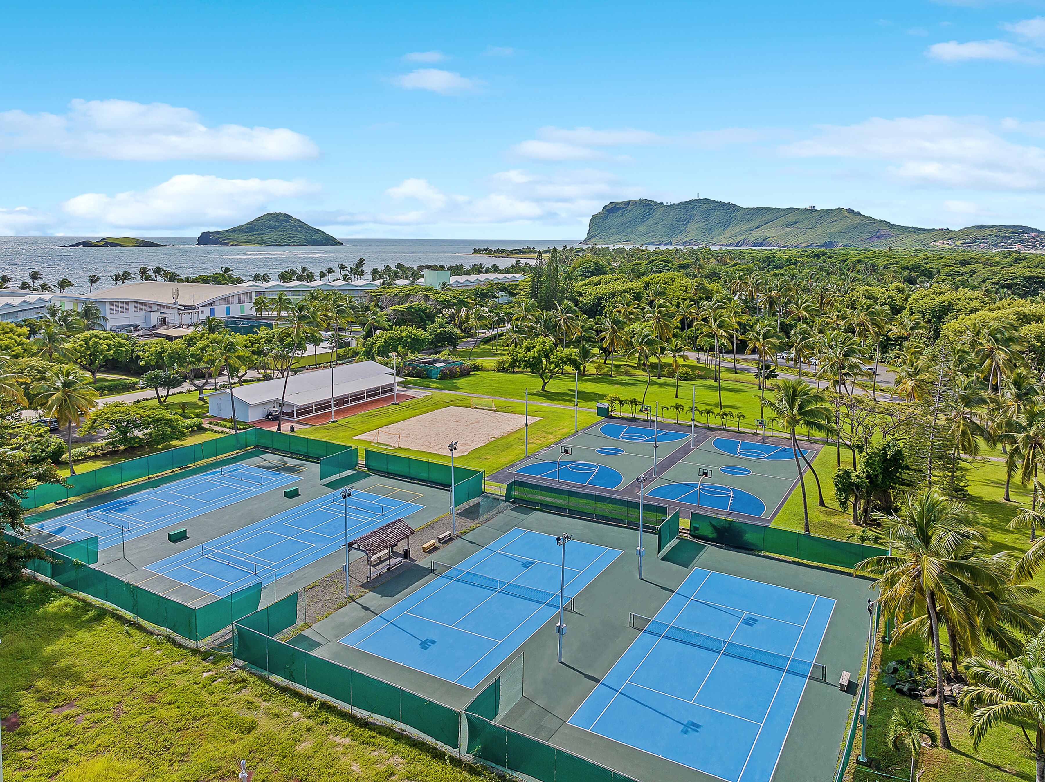 Een sportcomplex met verschillende tennisbanen, omringd door groen en uitzicht op zee en heuvels.