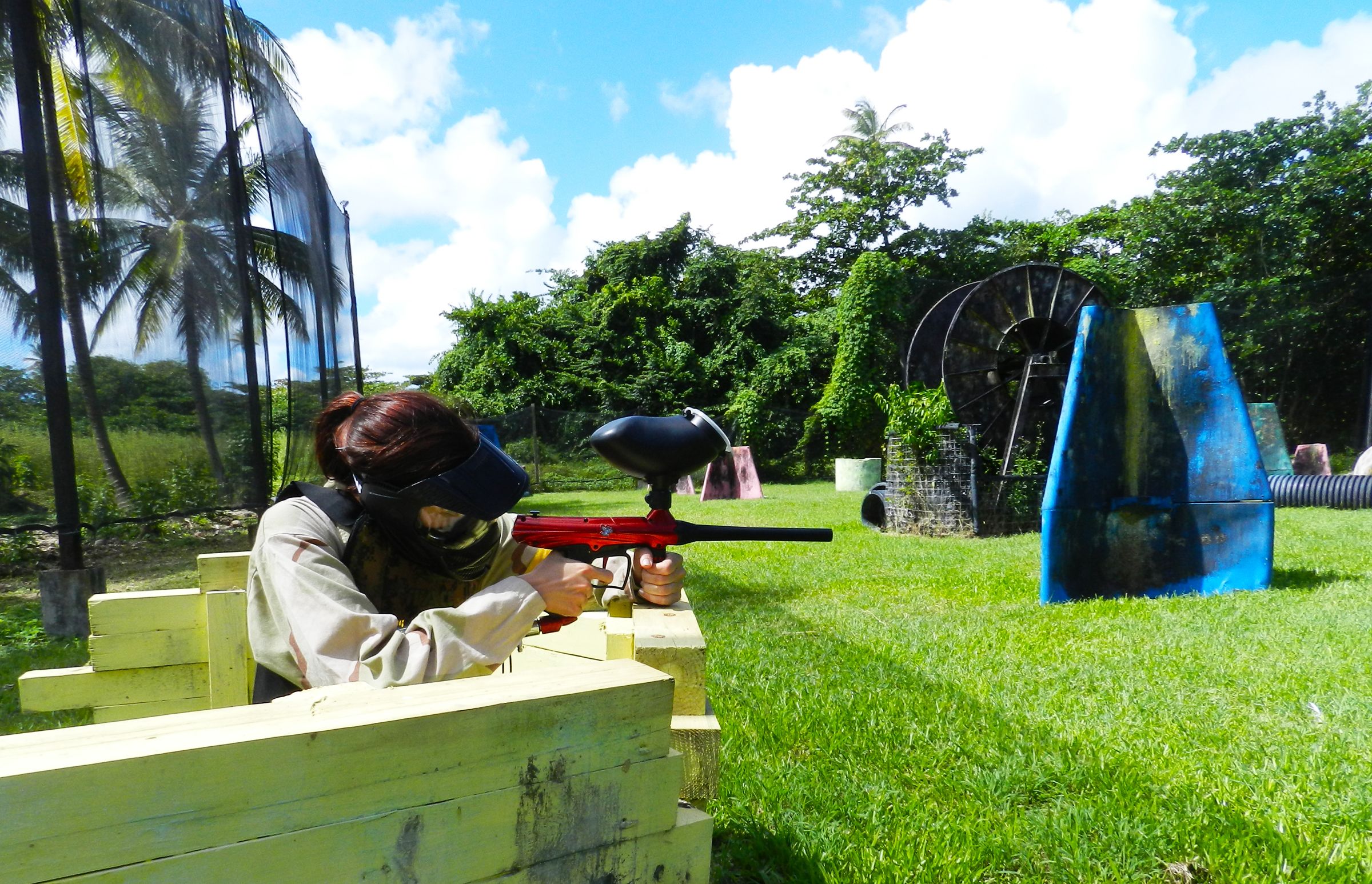 Una persona apuntando con un arma de paintball a un blanco en un entorno al aire libre con vegetación y cielo despejado.