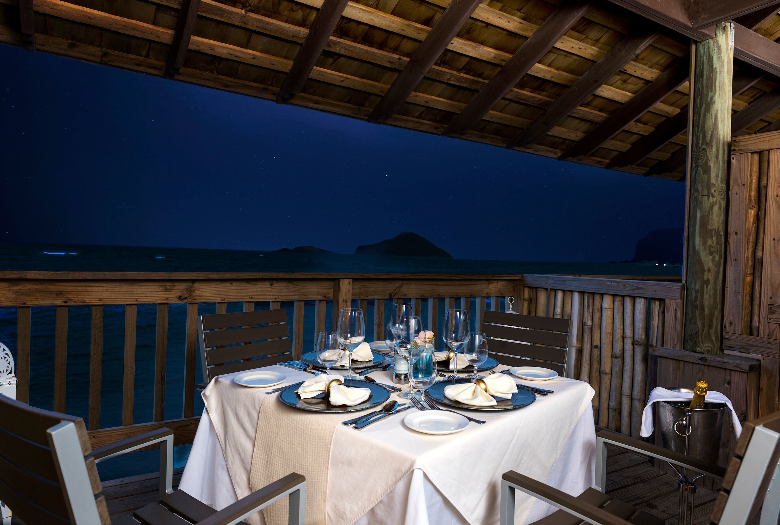 A table set for dinner on a wooden deck with a view of the ocean at night.