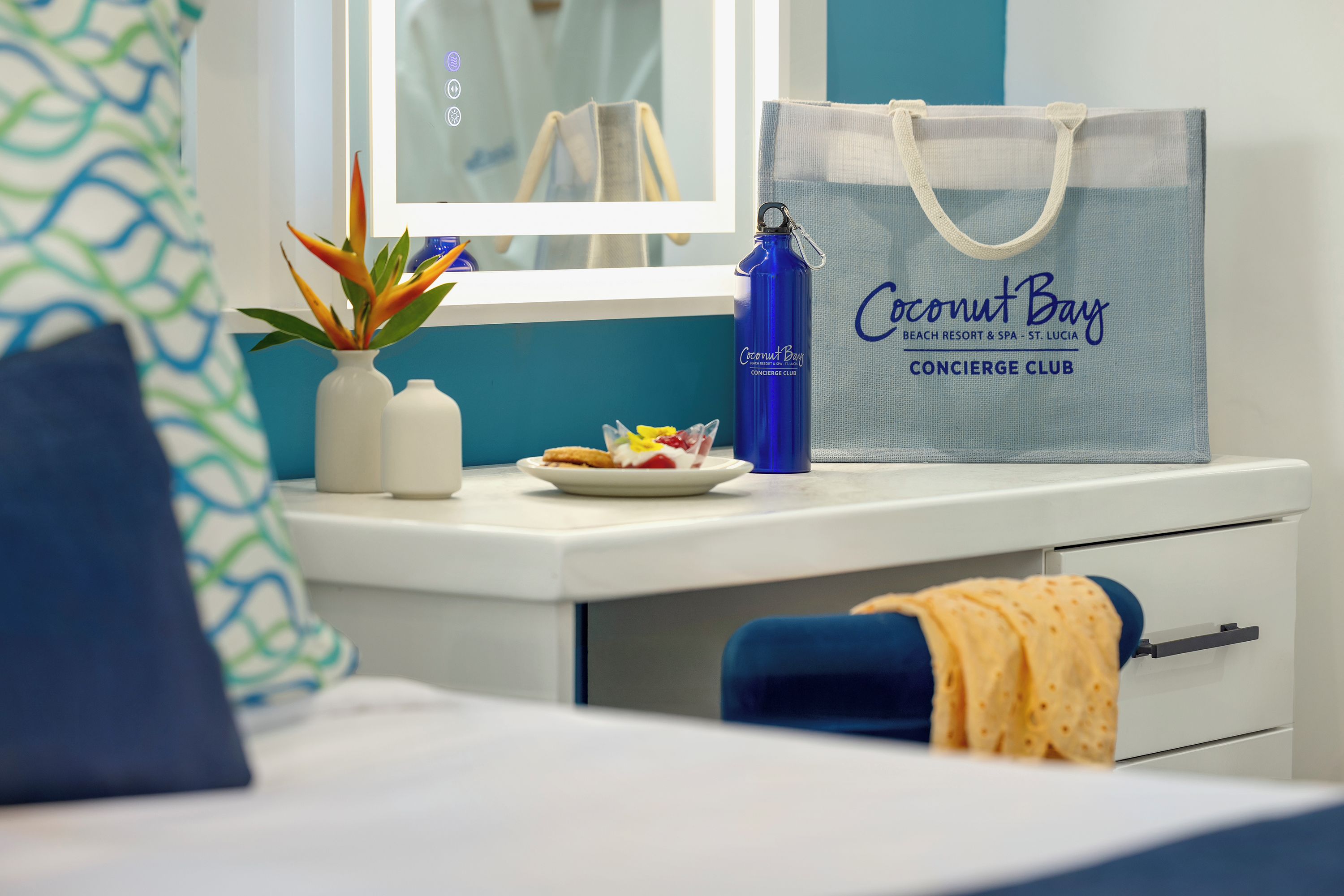 Una habitación de hotel con una cama, un escritorio con una botella de agua azul, una bolsa blanca y una planta pequeña.