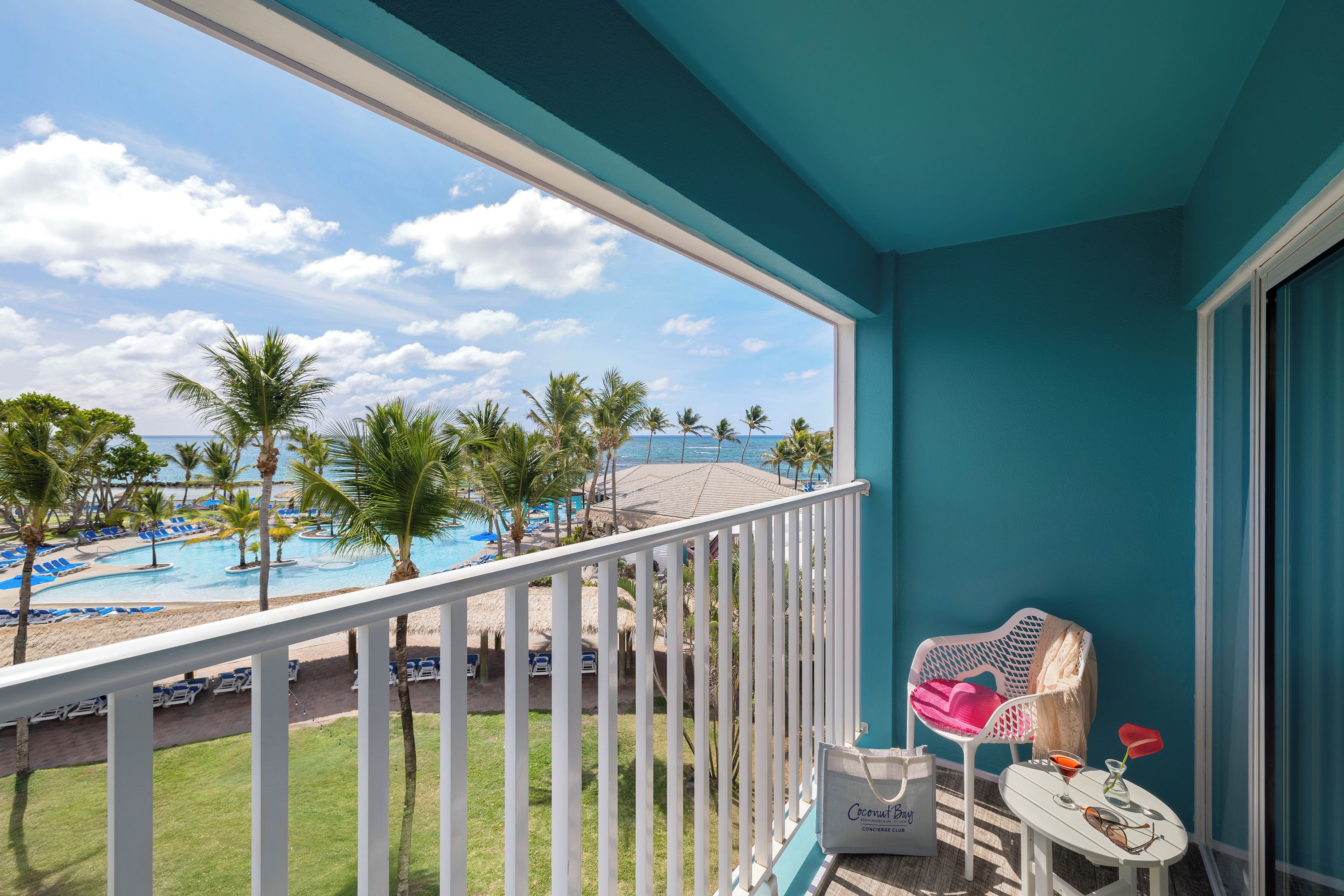 A balcony with a view of a pool and palm trees, a table with a bag and a drink, and a chair with a towel.