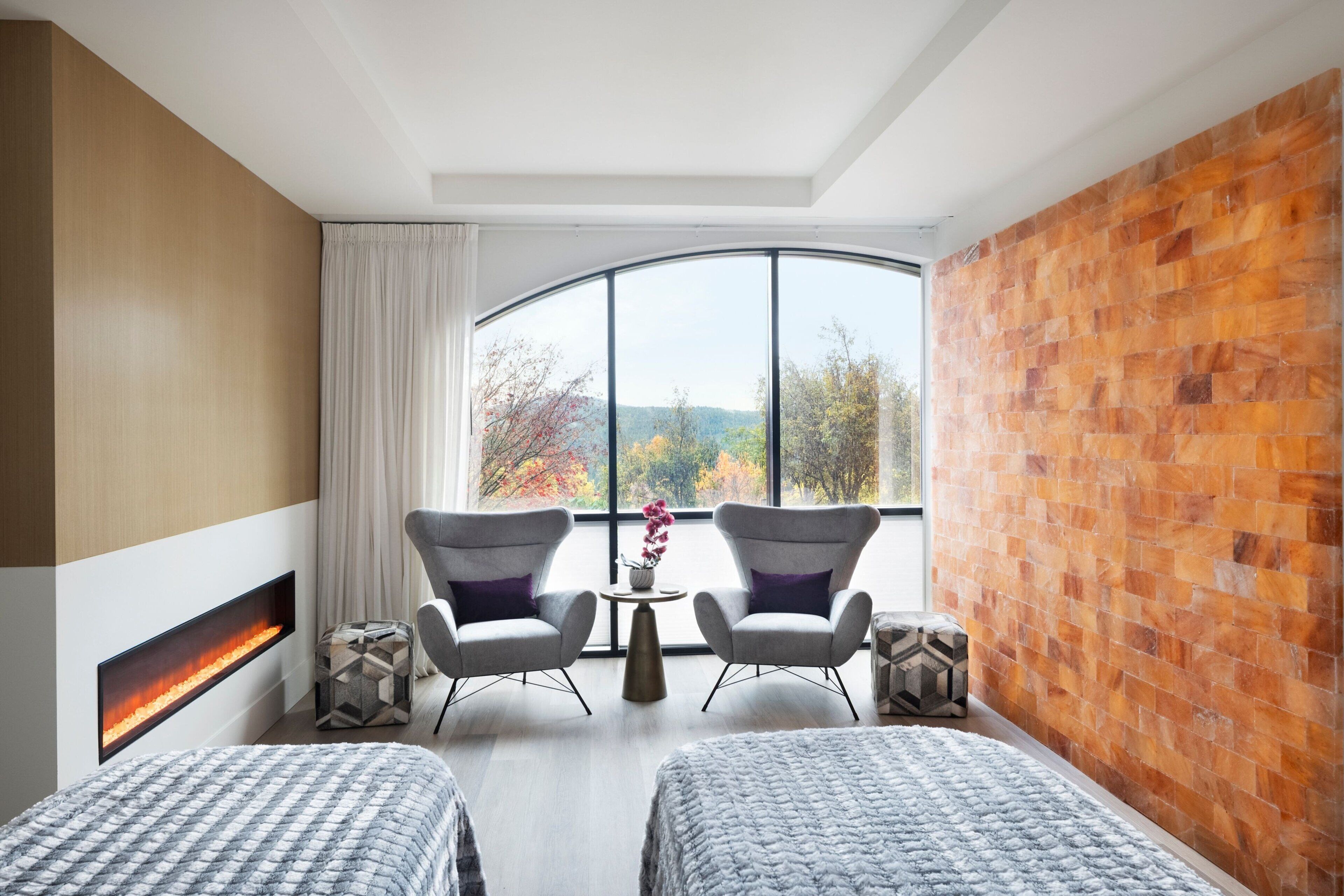 A hotel room with two beds, a fireplace, a large window with a view of trees, and a brick accent wall.