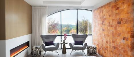 A hotel room with two beds, a fireplace, a large window with a view of trees, and a brick accent wall.