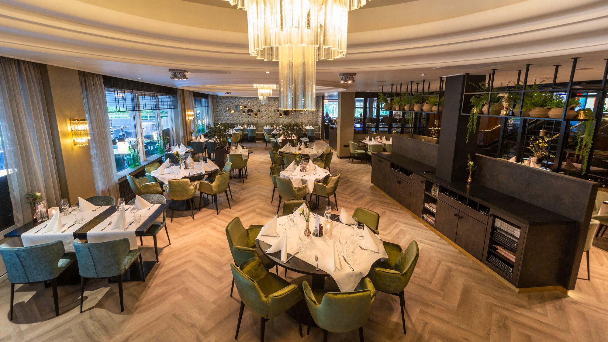 Een restaurant met ronde tafels, groene stoelen en een bar met plantenrekken.