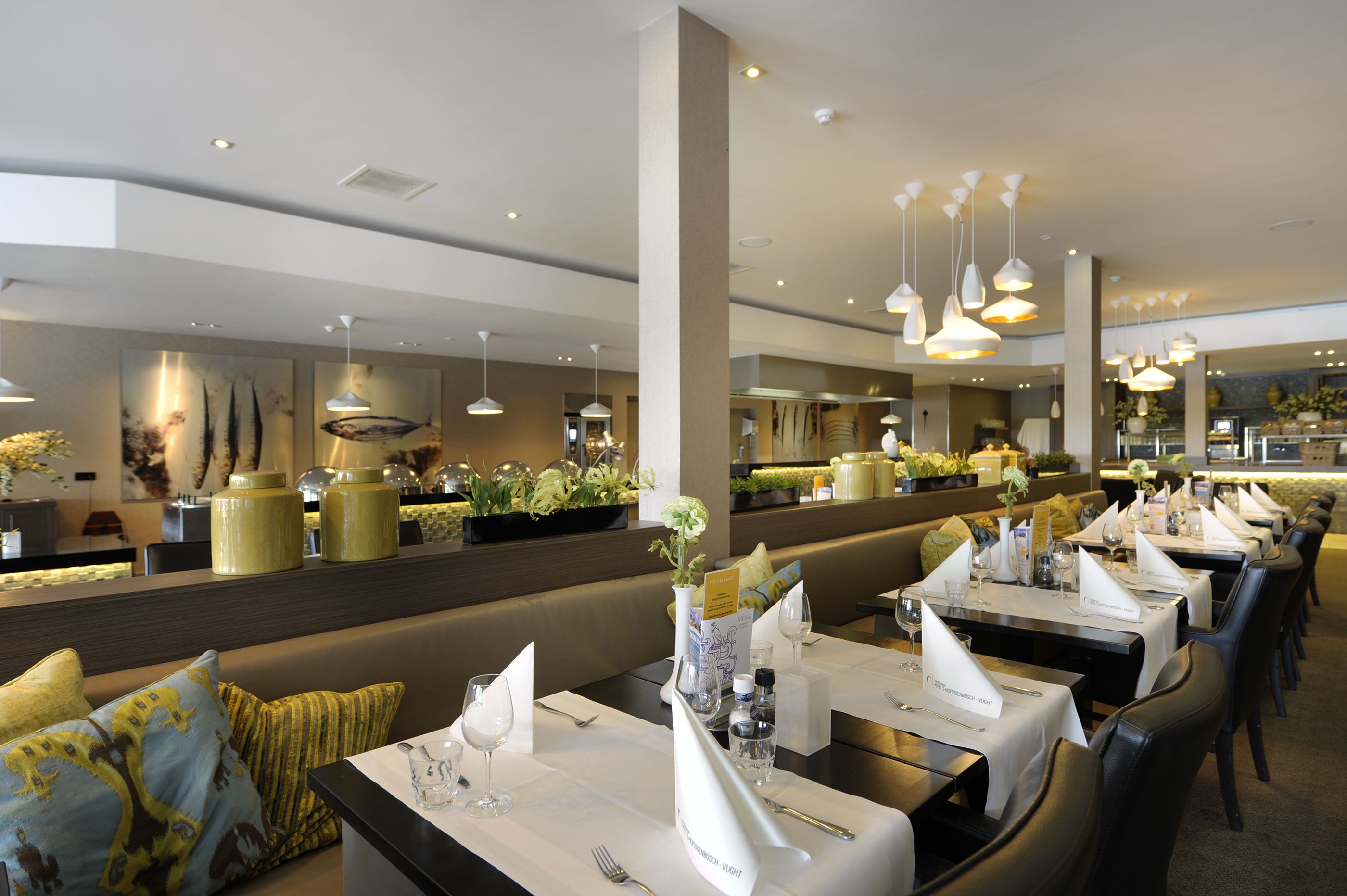 A restaurant with multiple dining tables set with white tablecloths, silverware, and glassware. There are hanging pendant lights above the tables, and a counter with a display of food items and plants.