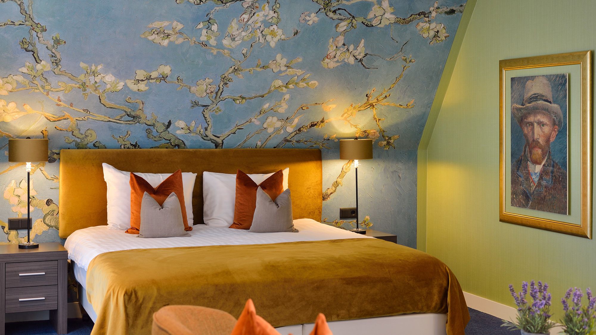 A bedroom with a large bed, two bedside tables, a nightstand, a wall-mounted lamp, and a framed portrait.