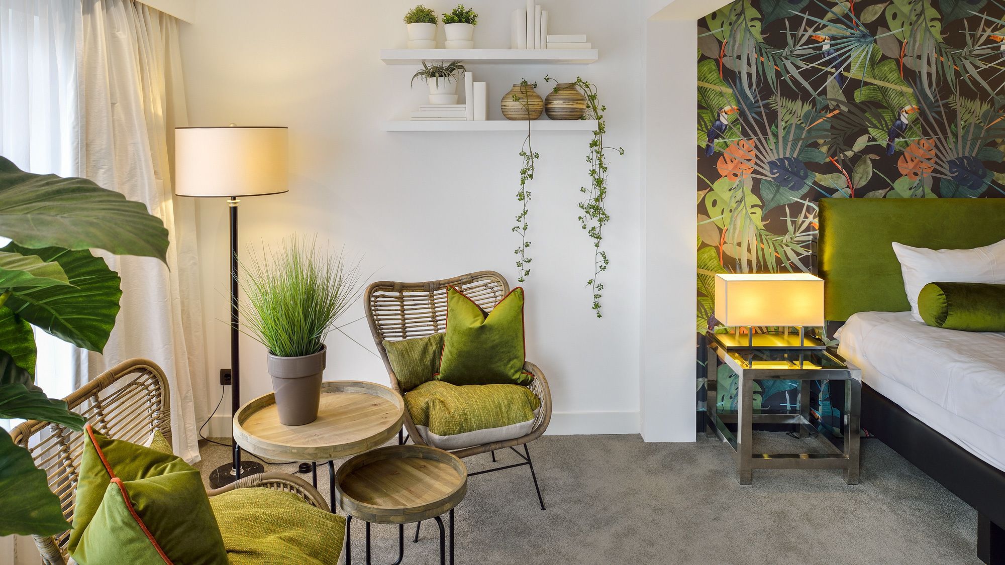A hotel room with a bed, a chair, a small table, a lamp, and a wall with a tropical plant mural.