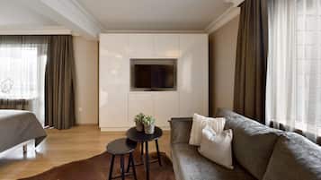 A modern hotel room with a large flat-screen TV mounted on a wall, a sofa with decorative pillows, a small round table with a plant, and a bed with a grey headboard.