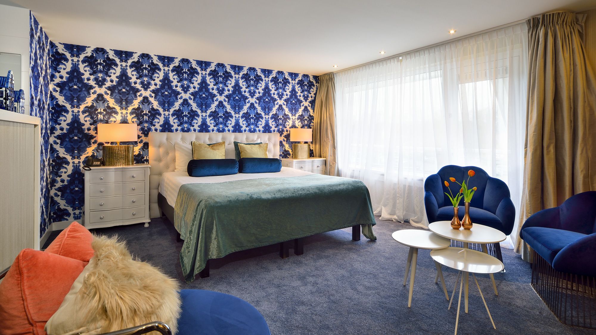 A hotel room with a bed, a nightstand, a chair, a small table, and a window with curtains.