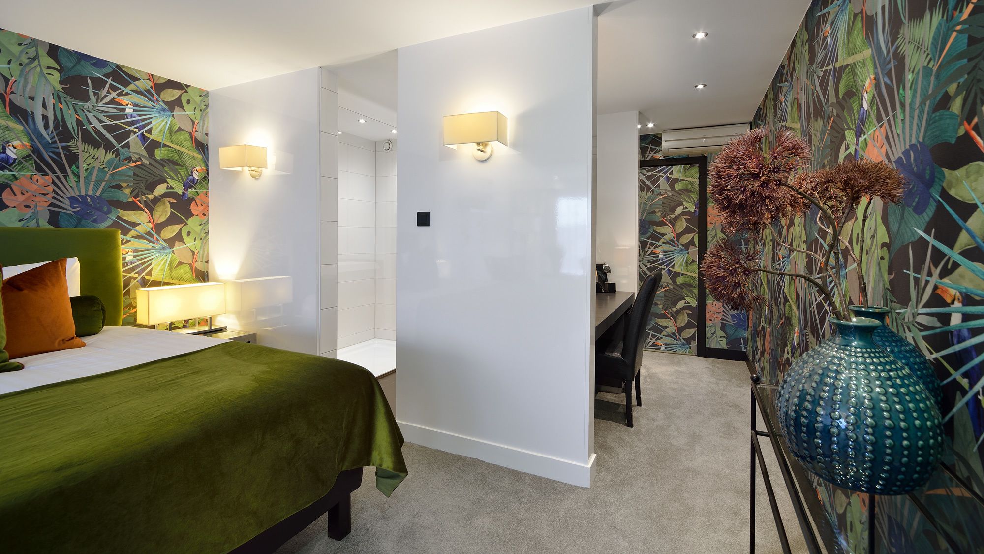 A modern bedroom with a green bed, two bedside lamps, a large blue vase with dried flowers, and a vibrant tropical wallpaper.