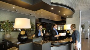 A reception desk with two staff members, a large lamp, and a potted plant.