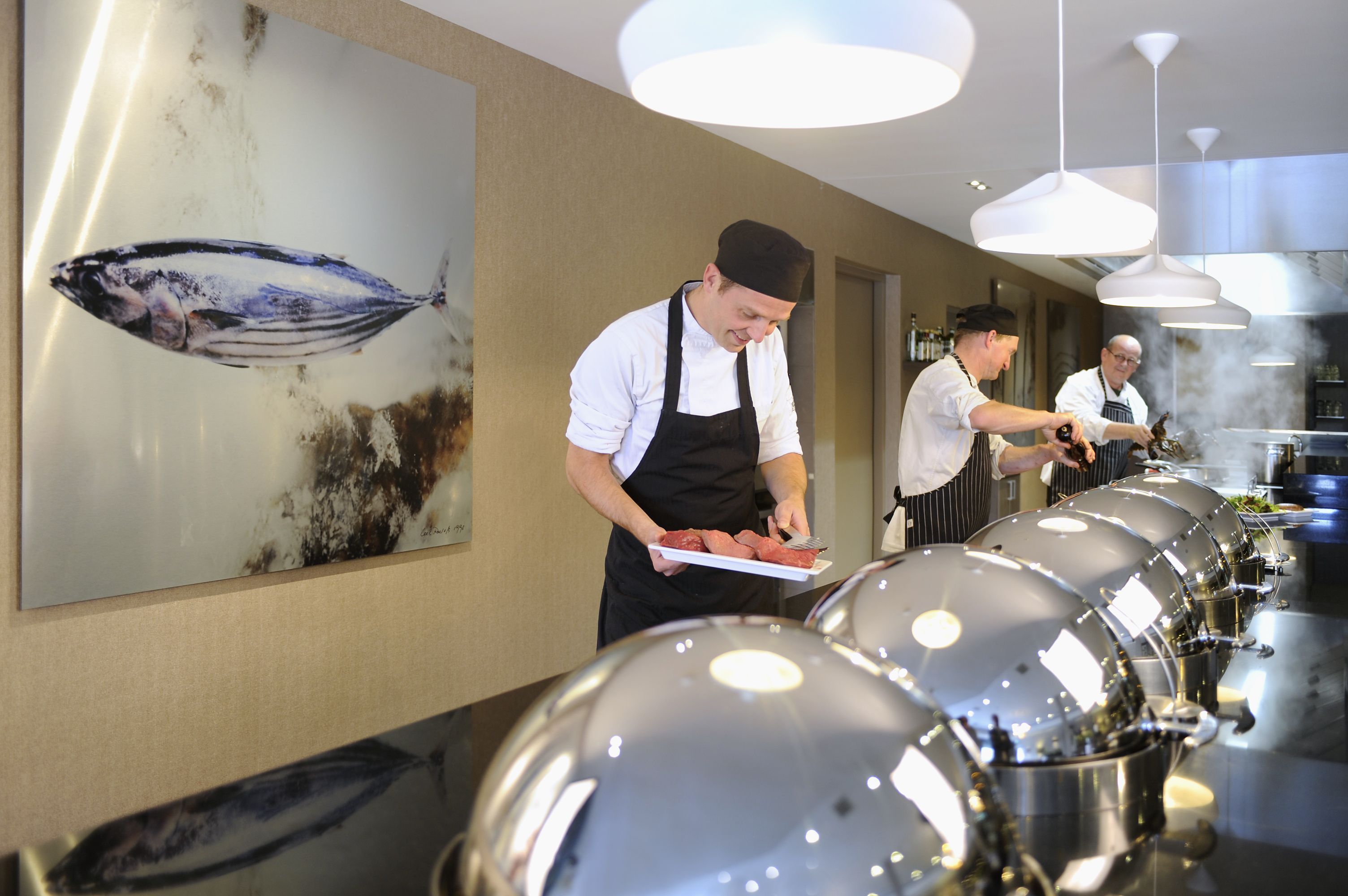 Een keuken met koks die eten bereiden, een groot schilderij van een vis aan de muur, en moderne hanglampen.