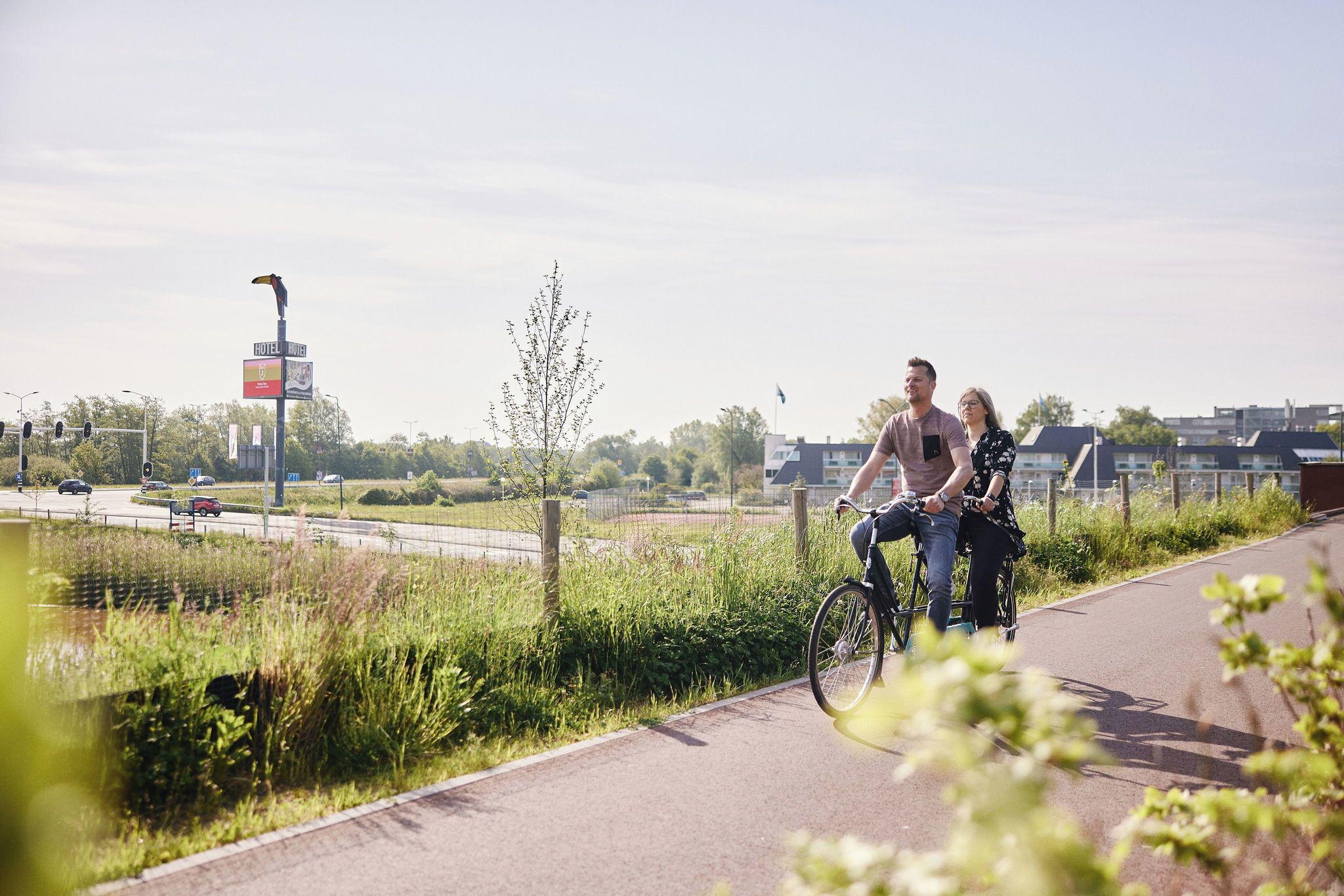 A couple riding a bicycle on a paved path with a hotel sign in the background.