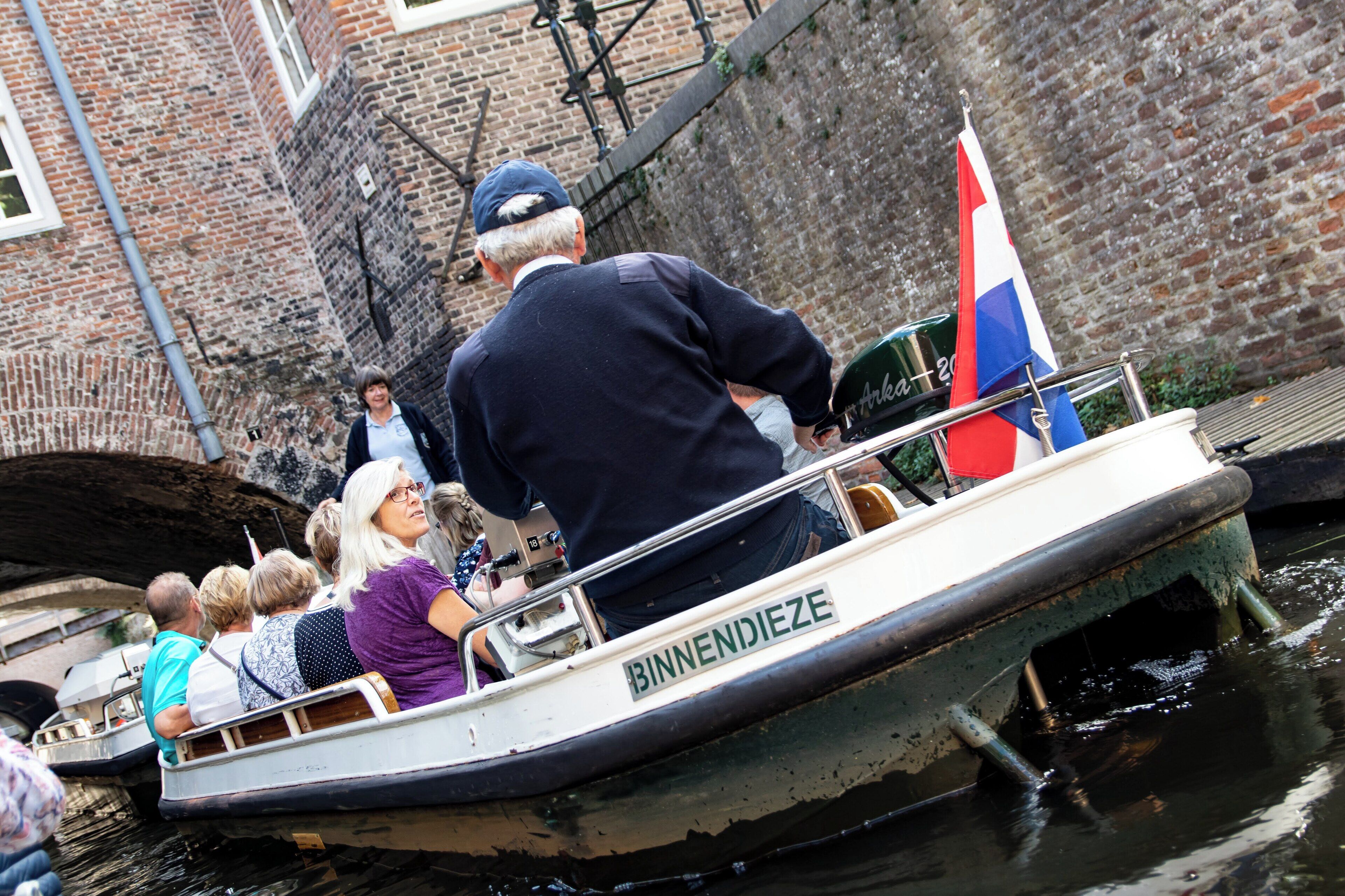 A narrow canal boat with passengers, a flag, and a brick wall in the background.