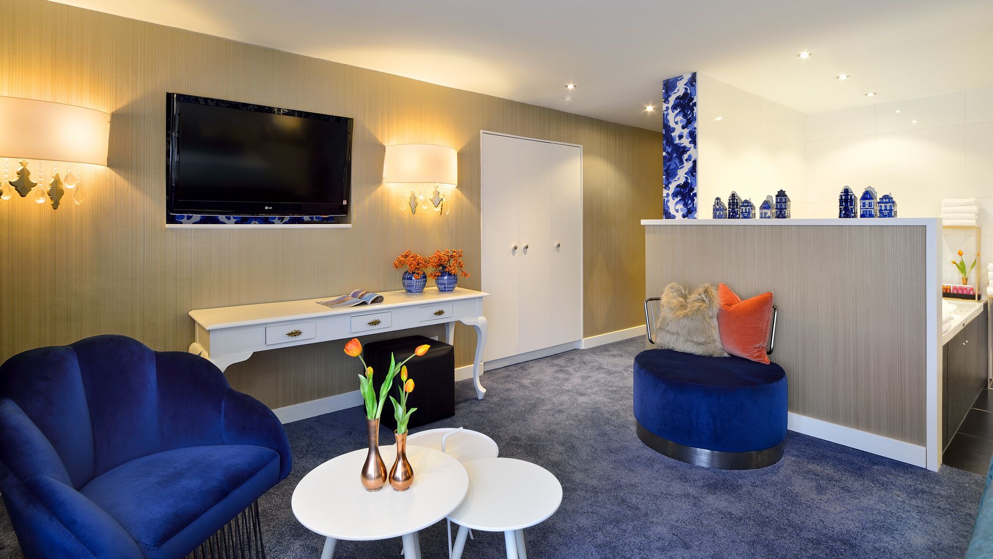 Een moderne hotelkamer met een flatscreen televisie, een witte console tafel met decoratieve objecten, een blauwe fauteuil, een ronde salontafel en een blauwe accentstoel.