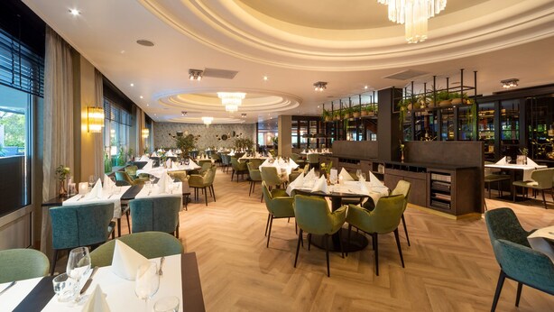 A spacious restaurant with multiple dining tables and chairs, a bar area with shelves, and large windows allowing natural light.