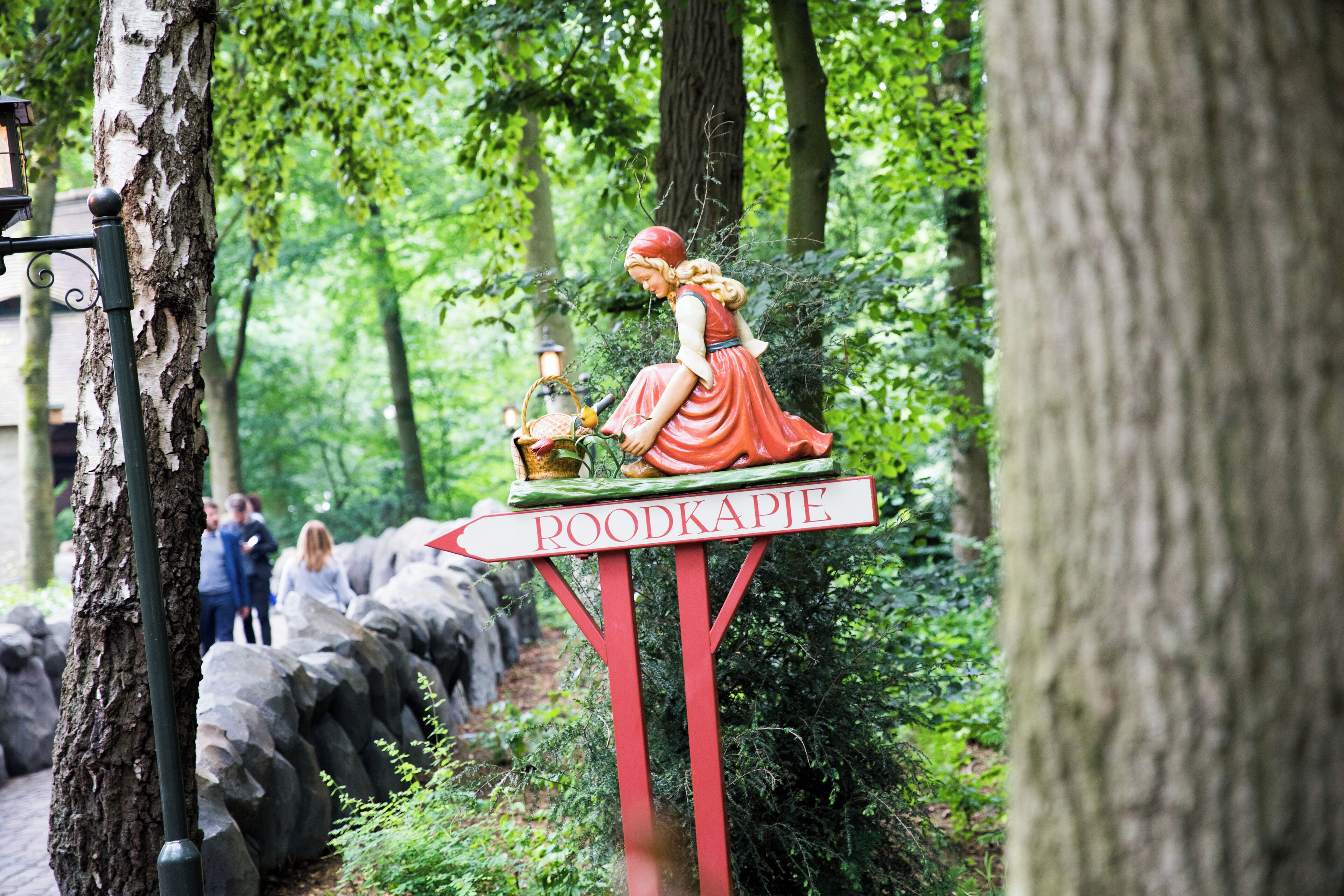 Ein Wegweiser mit einer Frauenfigur und dem Wort 'ROODKAPJE', umgeben von Bäumen und Felsen.