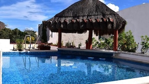 Piscina com estrutura de telhado de palha, rodeada por plantas e um céu azul claro.