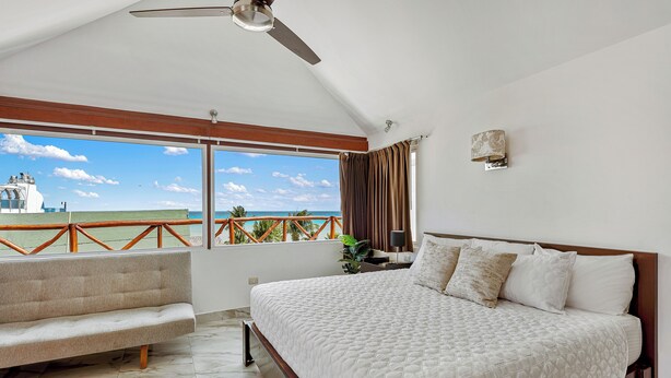 Quarto com uma cama grande, um banquinho, um ventilador de teto, uma mesinha de cabeceira e vista para o oceano.