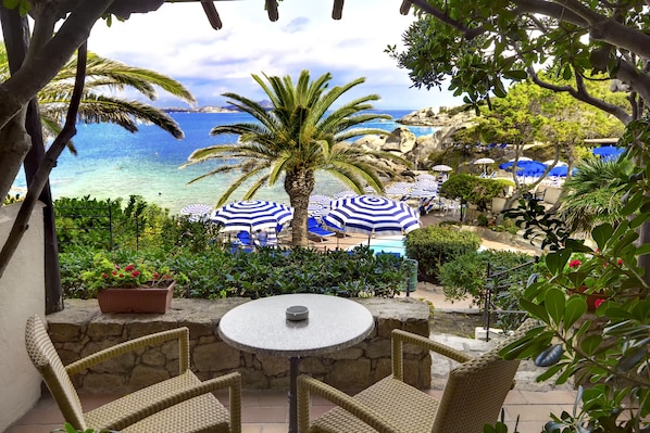 Un patio con sillas de mimbre y una mesa, con vistas a una zona de piscina con sombrillas azules y blancas a rayas, una palmera y vista al mar.