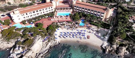 An aerial view of a coastal resort with a large pool, beach area with sun loungers, and a rocky shoreline.