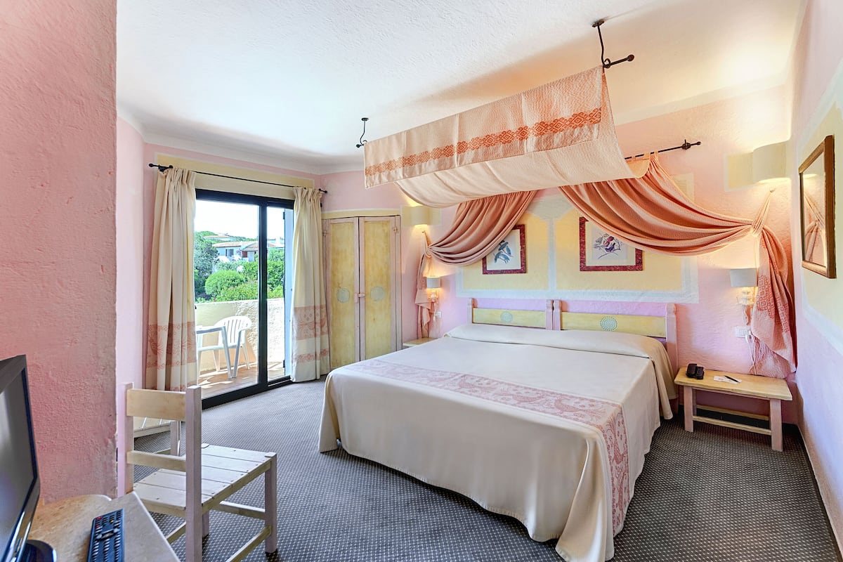 A hotel room with a bed, a chair, a desk with a remote control, and a view of the outdoors.