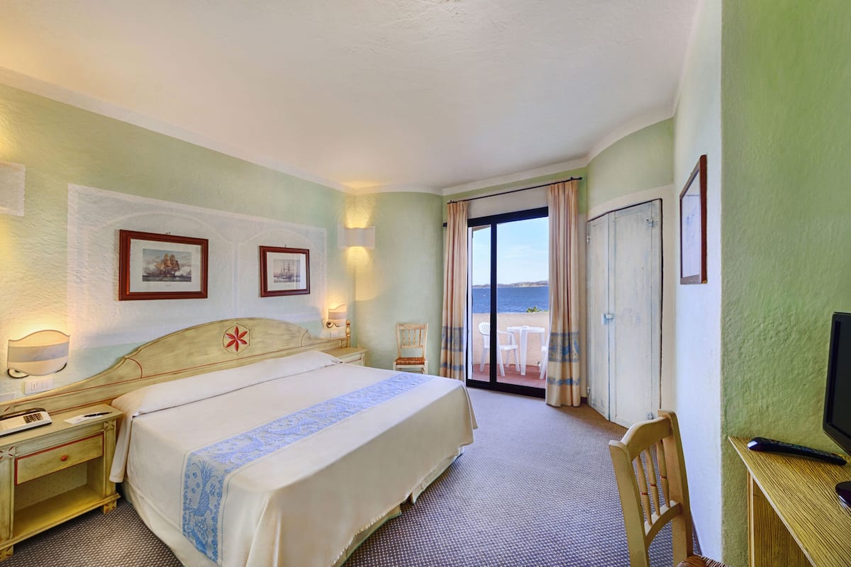 A hotel room with a bed, a desk with a computer, a chair, a nightstand, and a view of the beach through the door.