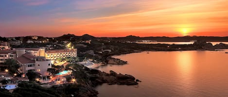 Un complejo turístico costero con un gran edificio, una piscina y vistas al atardecer sobre el agua.