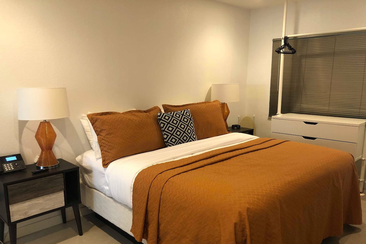 A neatly made bed with a brown blanket, two bedside lamps, a bedside table with a lamp, and a window with blinds.