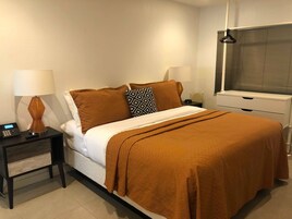 Una cama bien hecha con una manta marrón, dos lámparas de noche, una mesita de noche con lámpara, y una ventana con persianas.