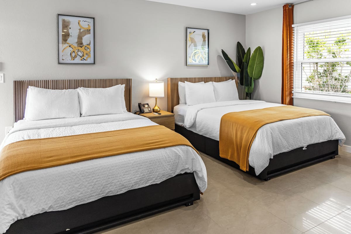A hotel room with two beds, a nightstand with a lamp, and a large window with blinds.