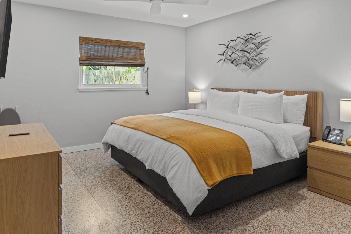 A bedroom with a bed, bedside tables, a desk, a window with blinds, and a wall-mounted decorative piece.