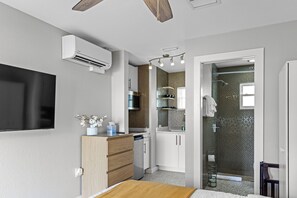 Un dormitorio compacto con cama, mesita de noche y un escritorio pequeño con silla. Cuenta con un armario empotrado y una zona de ducha con puerta de cristal.