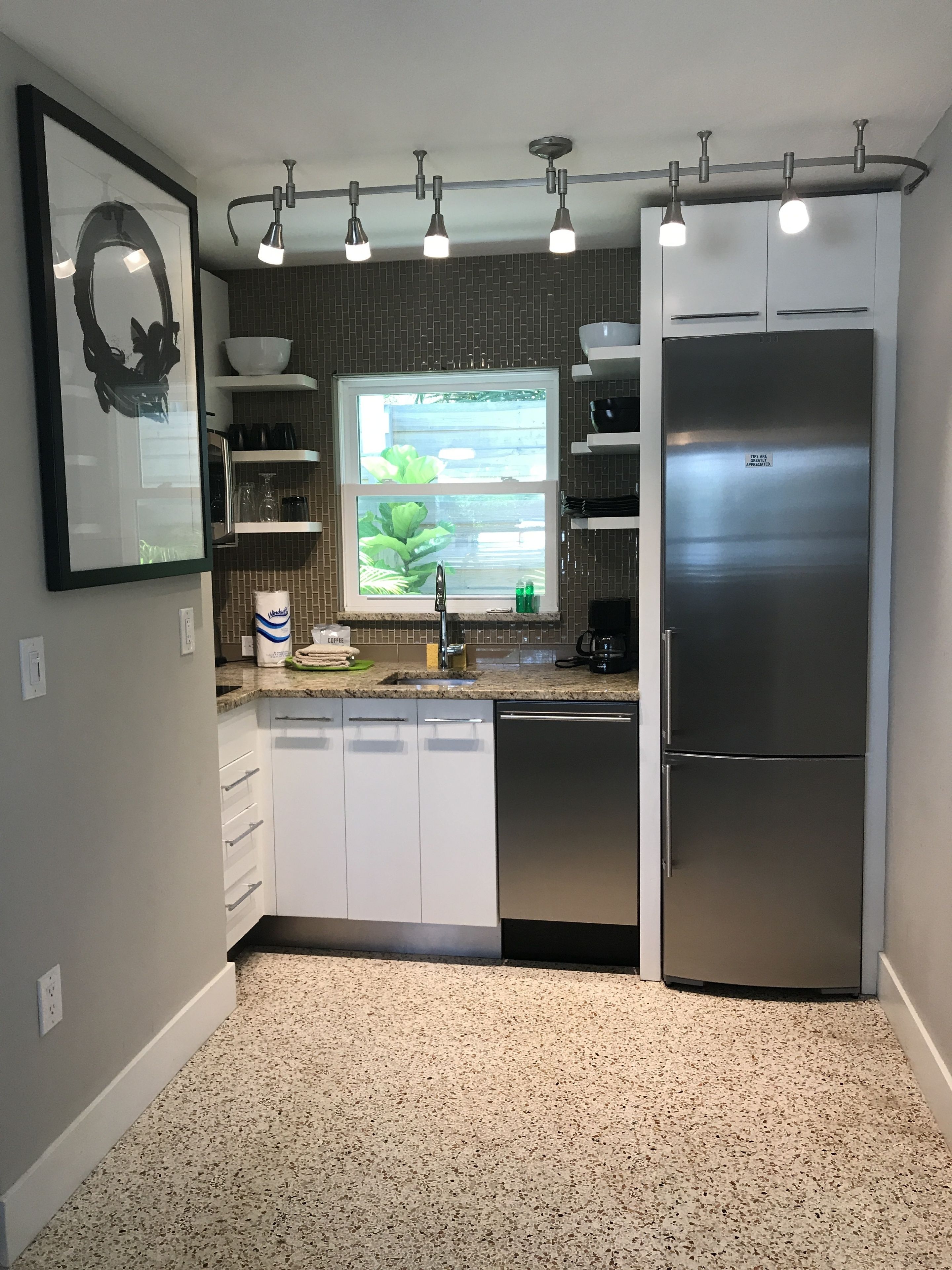 A modern kitchen with a stainless steel refrigerator, a window with a view of greenery, and a countertop with various kitchen items.