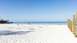Una playa con arena blanca, sillas de madera y un cielo azul despejado.