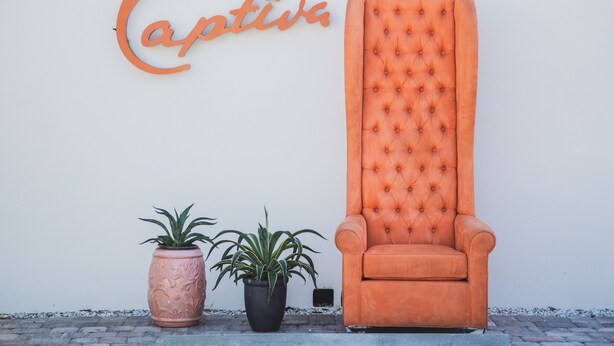 Un sillón naranja grande frente a una pared blanca con un letrero 'Captiva'. Hay dos macetas con plantas a un lado de la silla.