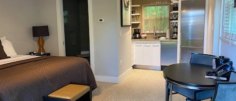 Habitación de hotel con cama, mesita de noche, lámpara y un pequeño banco. Se observa una kitchenette con refrigerador, estufa y microondas. También hay una mesa de comedor con sillas.