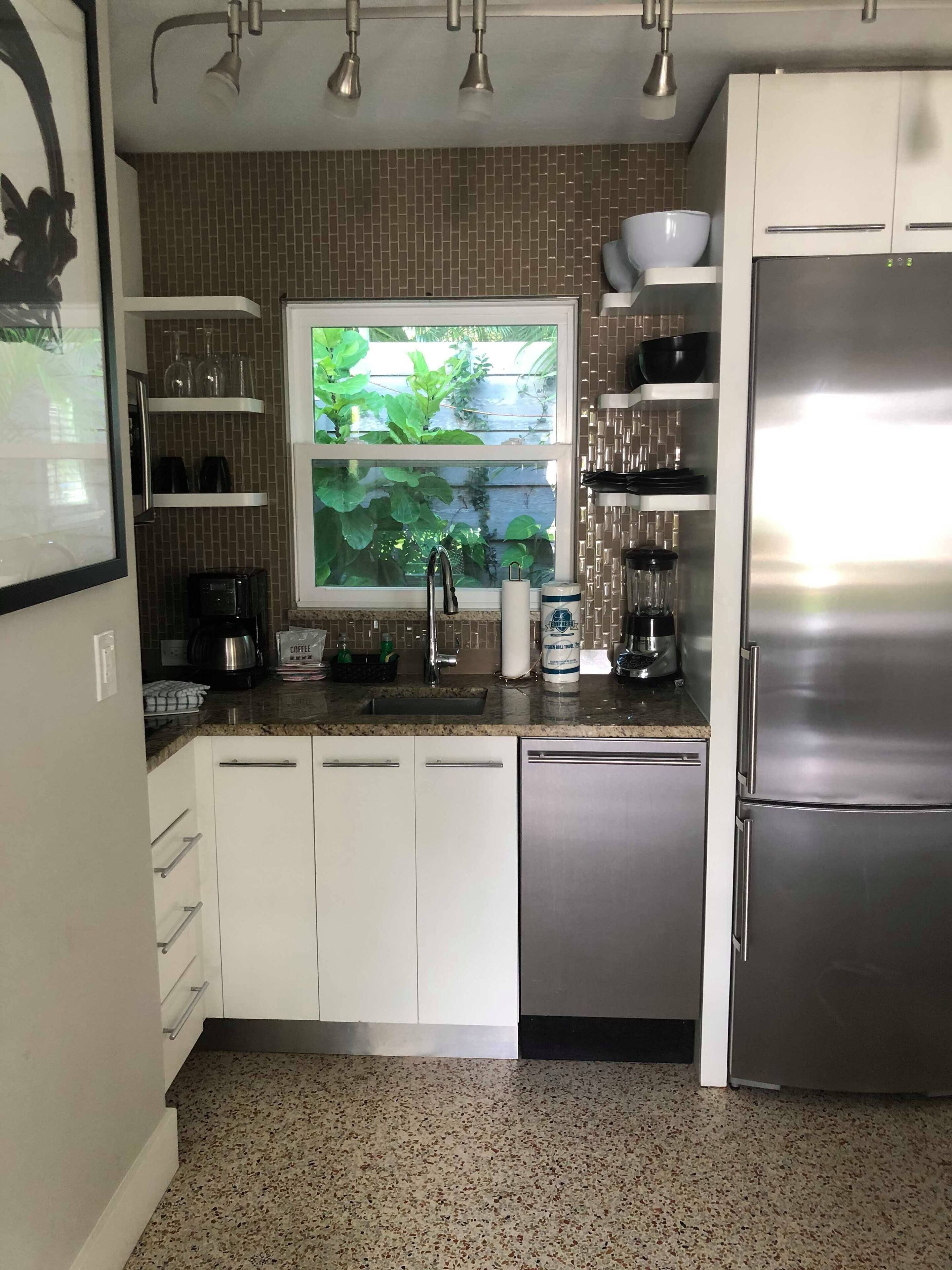 Una cocina moderna con armarios blancos, un refrigerador de acero inoxidable, una ventana con vistas a la vegetación y una cafetera.