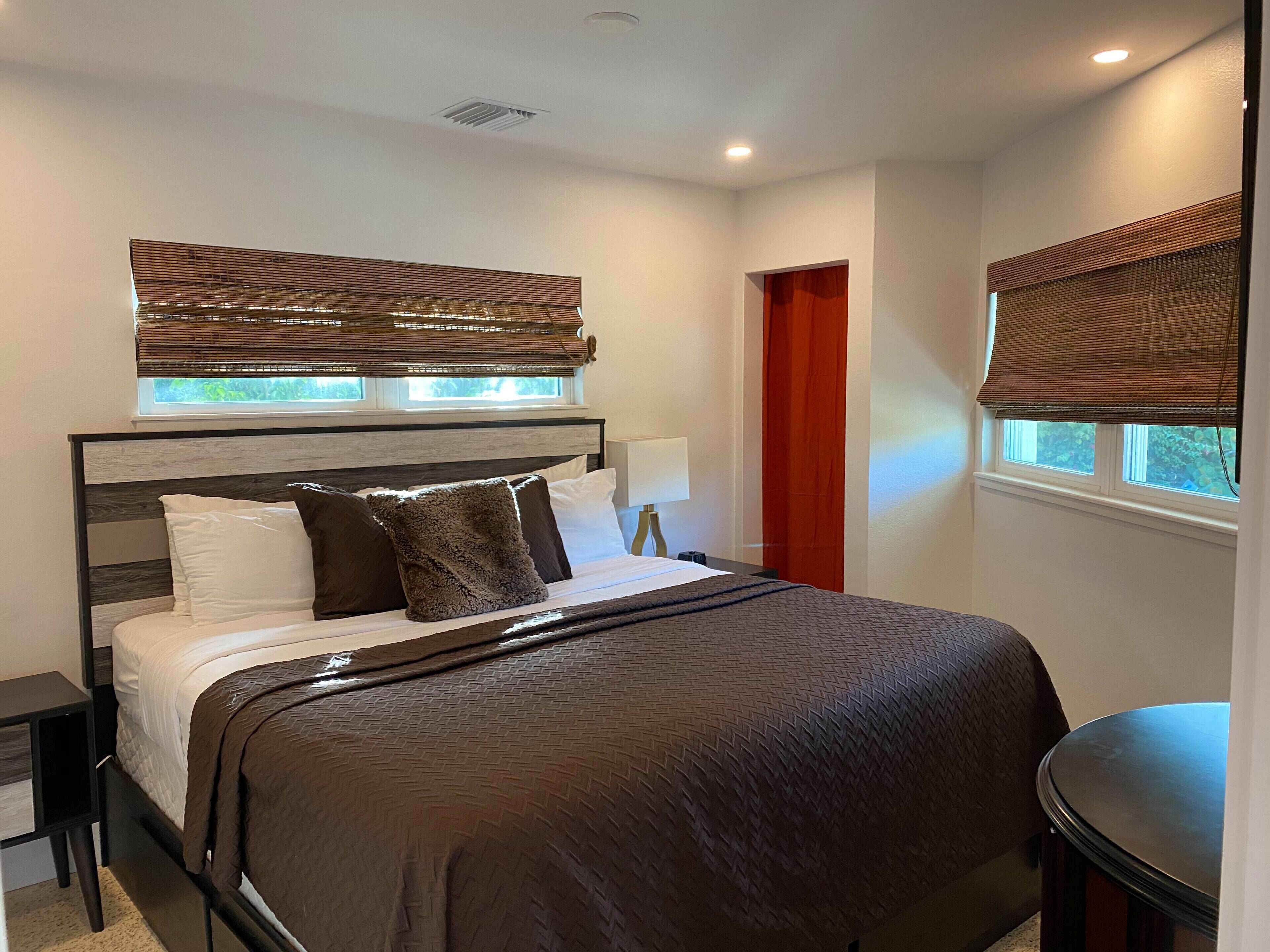 A modern bedroom with a large bed, a nightstand, a bedside table, a window with blinds, and a door.